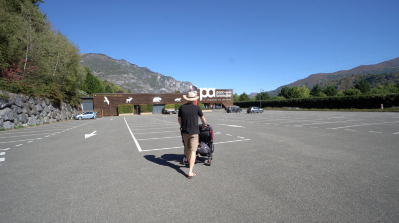 Man pushing stroller in large and mostly empty parking lot walking toward Parc Animalier Des Pyrénées entrance